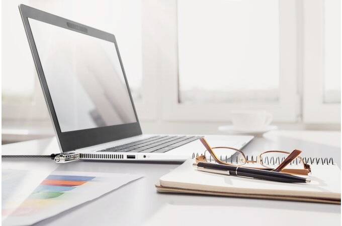 An open laptop, a pair of glasses, and a notepad with a pen on a desk in front of a window letting in bright daylight.