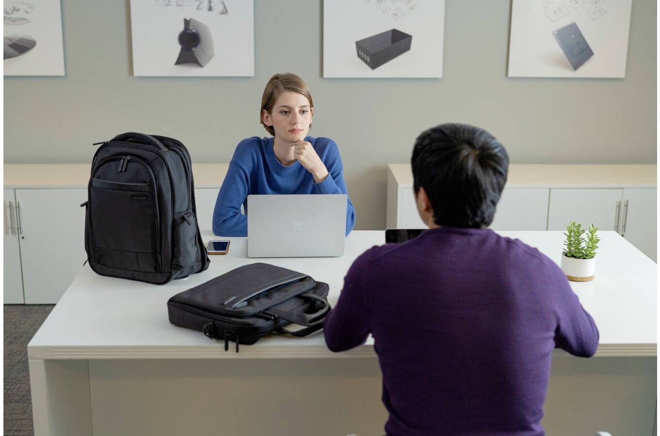 Two people are sitting at a desk in an office. One person is working on a laptop, while the other is listening. Bags are placed on the table.