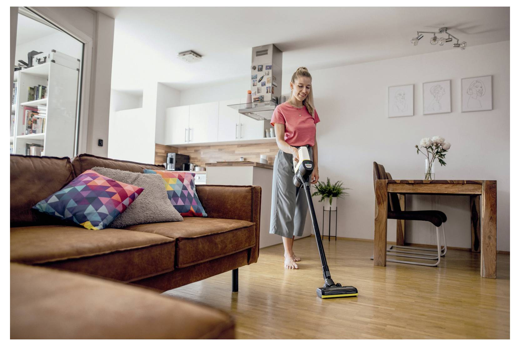A woman vacuums the living room floor of a modern, tidy apartment with wooden flooring and colorful pillows on the sofa.