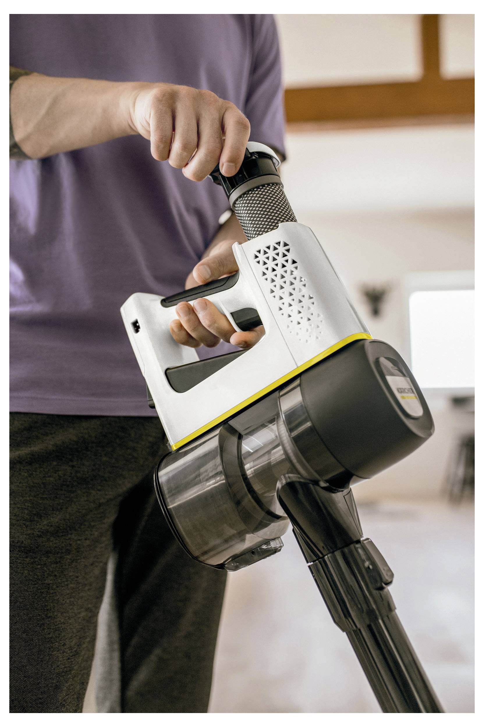 A person holds a modern cordless vacuum cleaner, focusing on the handle and upper section. The room appears bright and spacious.