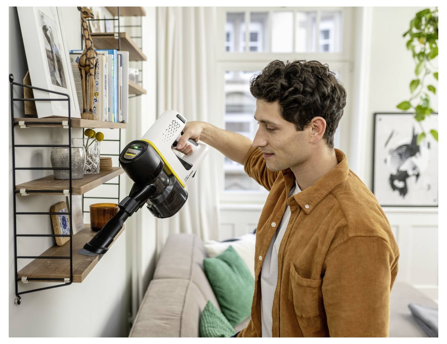 A person uses a handheld vacuum to clean a shelf, with books and decorative items, in a bright, modern living room.