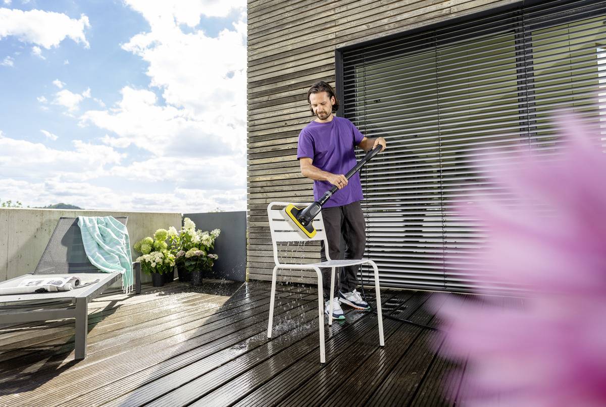 A person cleans a patio chair with a pressure washer on a sunny day. A towel hangs on a chair and plants are in the background.