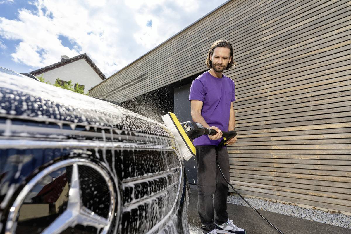 A person in a purple shirt is cleaning a black car with a power washer in front of a modern wooden garage under a partly cloudy sky.