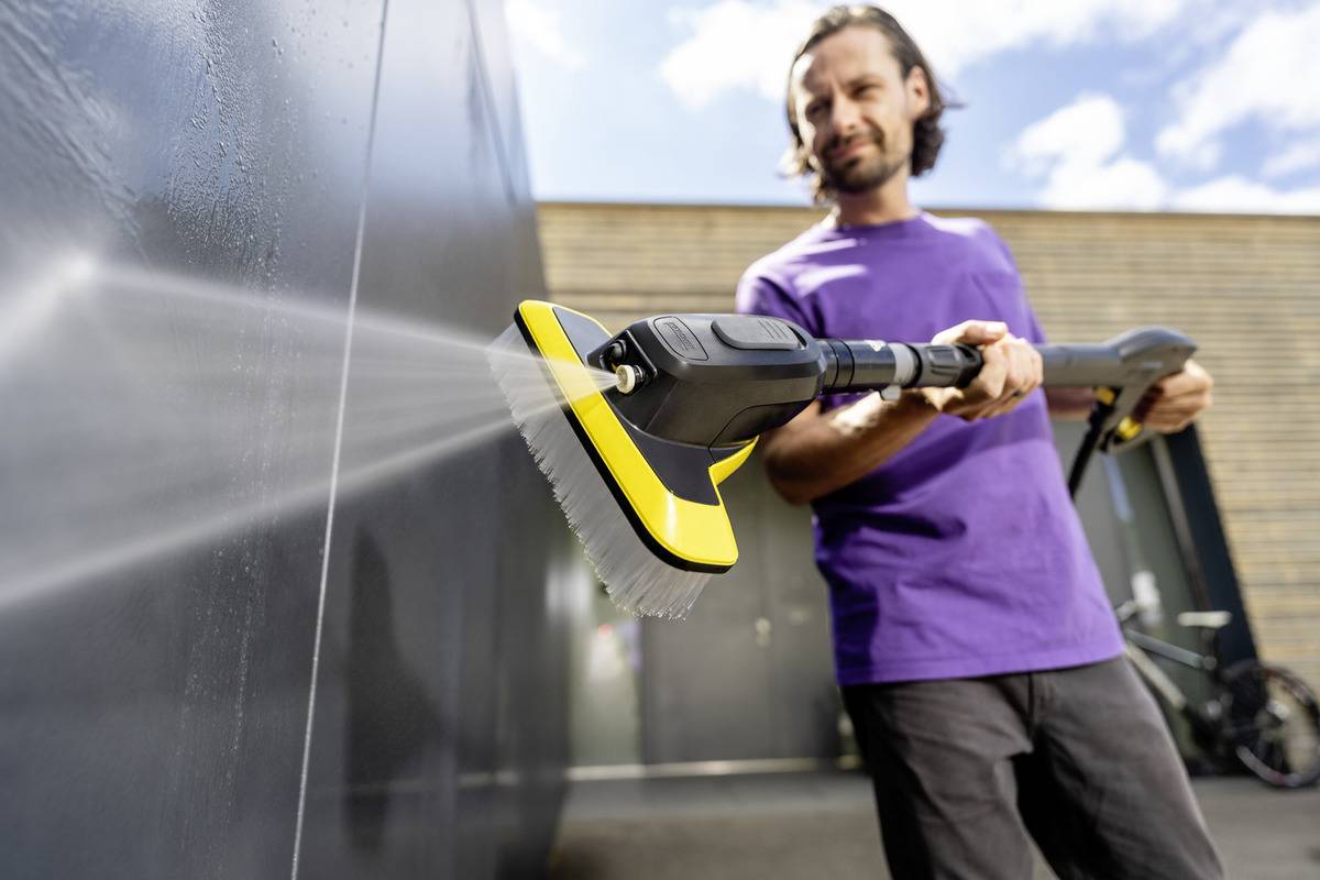 A person in a purple shirt uses a pressure washer with a brush attachment to clean an outdoor wall, focusing on removing dirt.