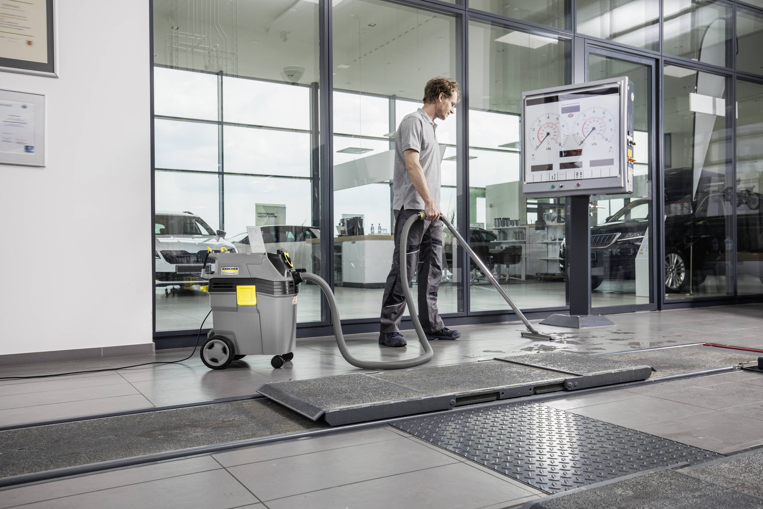 A man is operating a floor cleaning machine in a modern building with large windows. A display can be seen in the background.