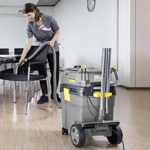 A woman is cleaning a room with a wooden floor and chairs using an industrial vacuum cleaner, indicating professional cleaning work.