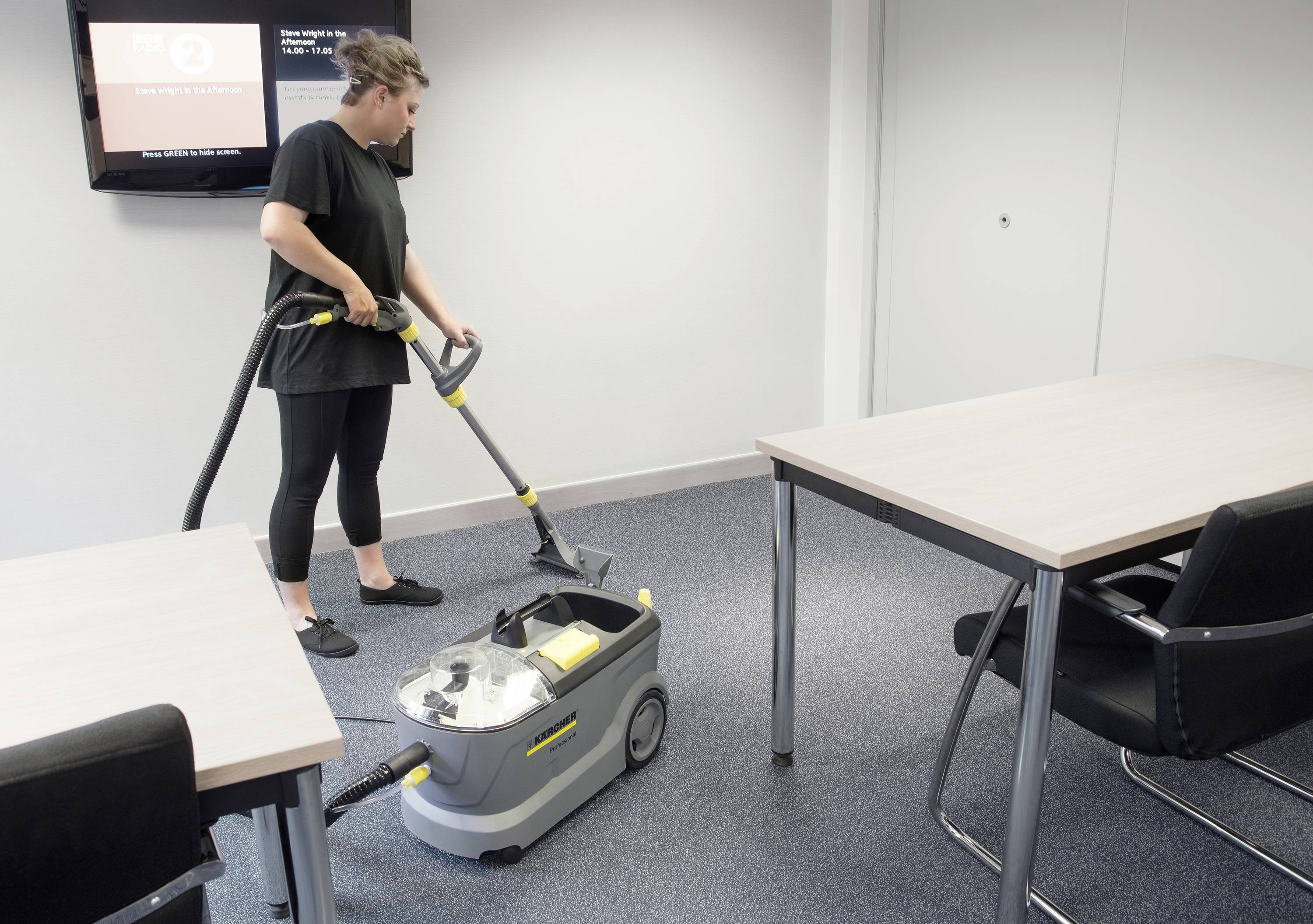 A person is vacuuming the floor with a carpet cleaner in a conference room with grey carpet and two tables with black chairs.