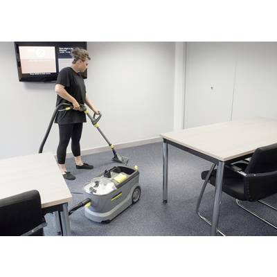 A person is vacuuming the floor with a carpet cleaner in a conference room with grey carpet and two tables with black chairs.