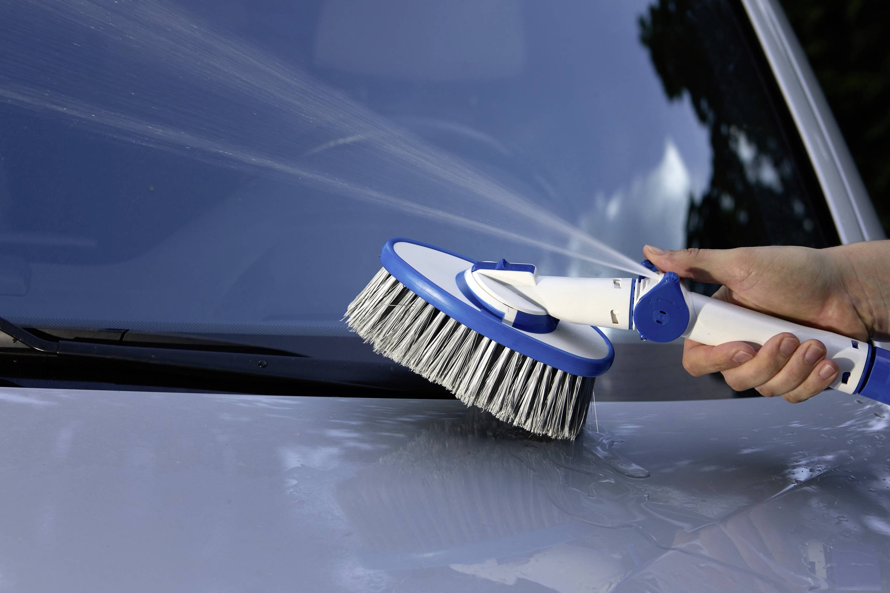 A person is cleaning a car with a brush that sprays water on the bonnet.