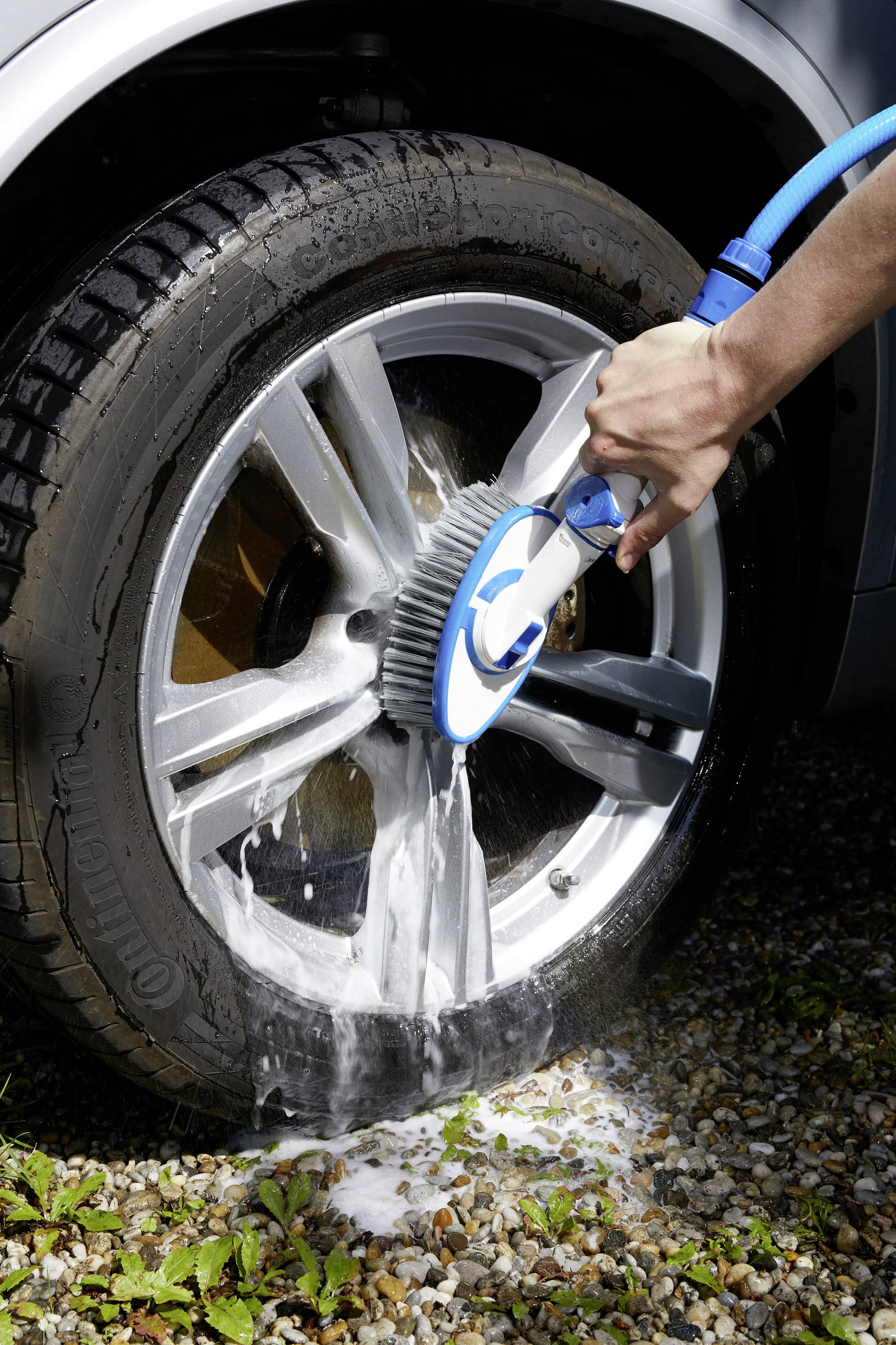 A person is cleaning a car tyre with a brush and water jet, foam visible on the tyre, stone floor in the background.