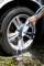 A person is cleaning a car tyre with a brush and water jet, foam visible on the tyre, stone floor in the background.