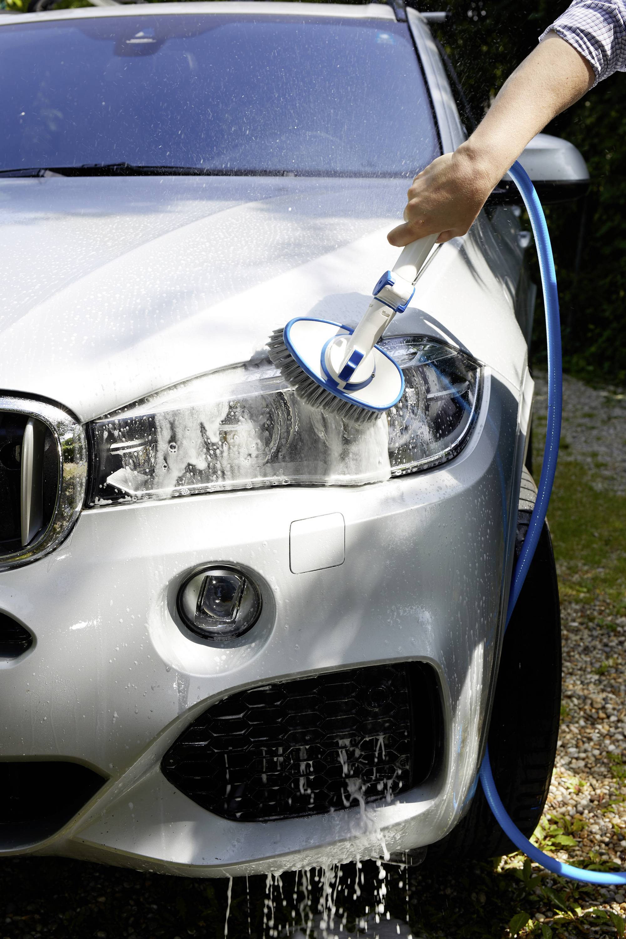 A person is cleaning a headlight of a silver car with a brush, with water running down the paintwork.
