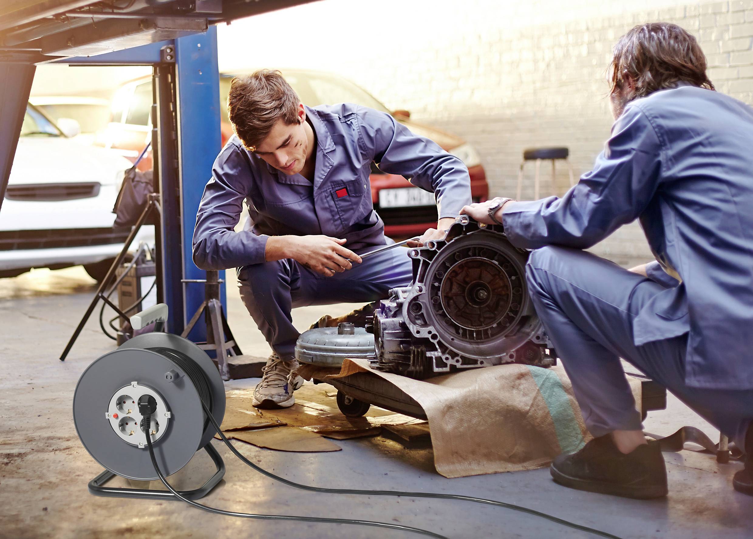 Two mechanics are repairing a car in a workshop, focused on an engine block. A white car is standing in the background.