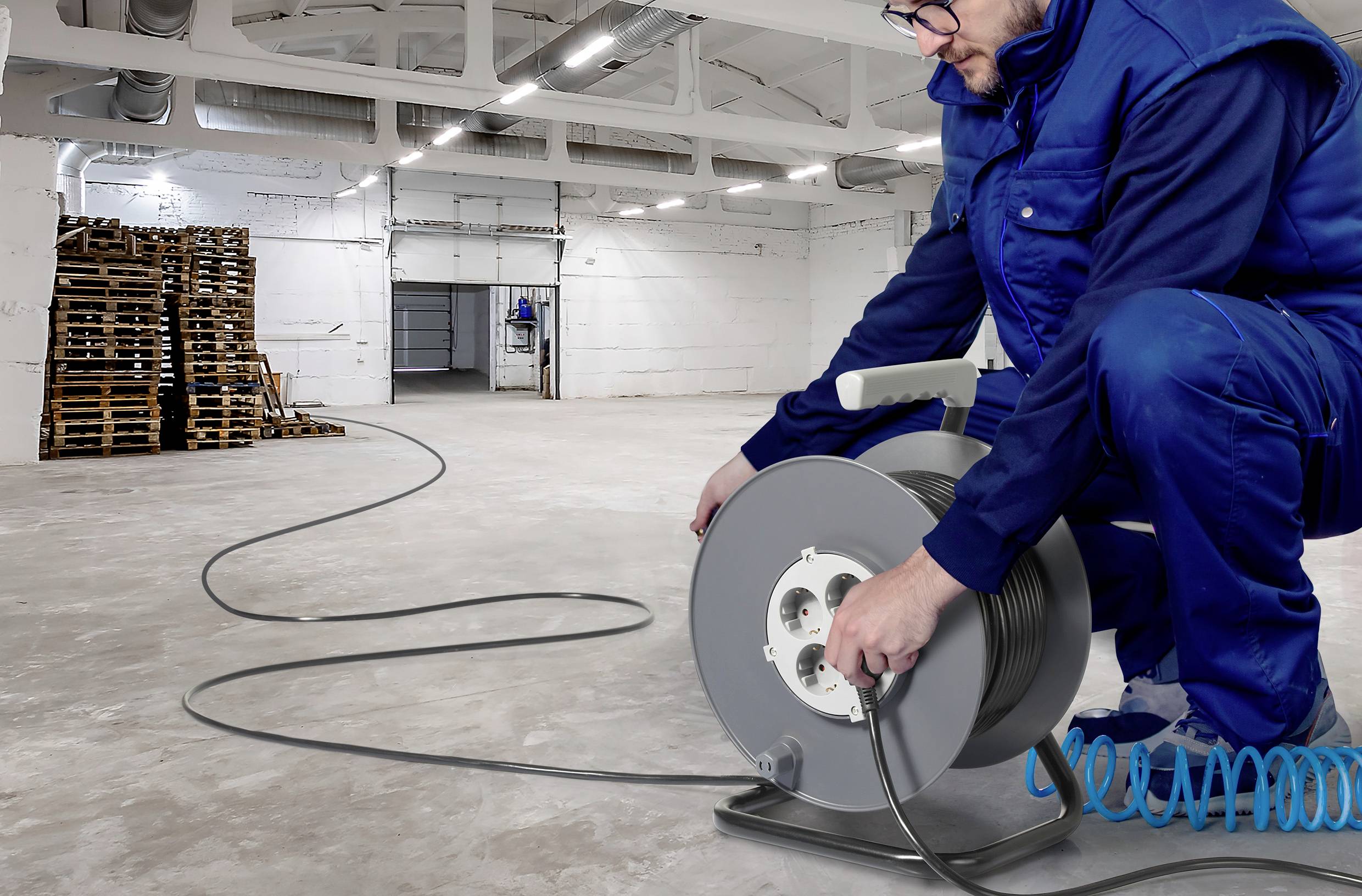 A man in workwear is winding a long cable onto a cable drum in a large, empty warehouse.