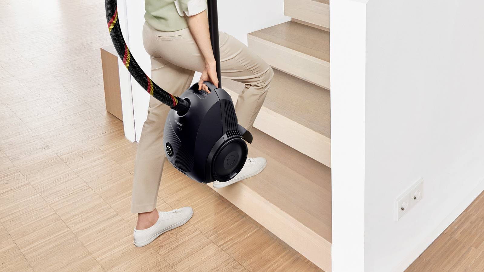 A person is walking up a staircase, carrying a black vacuum cleaner with a hose.