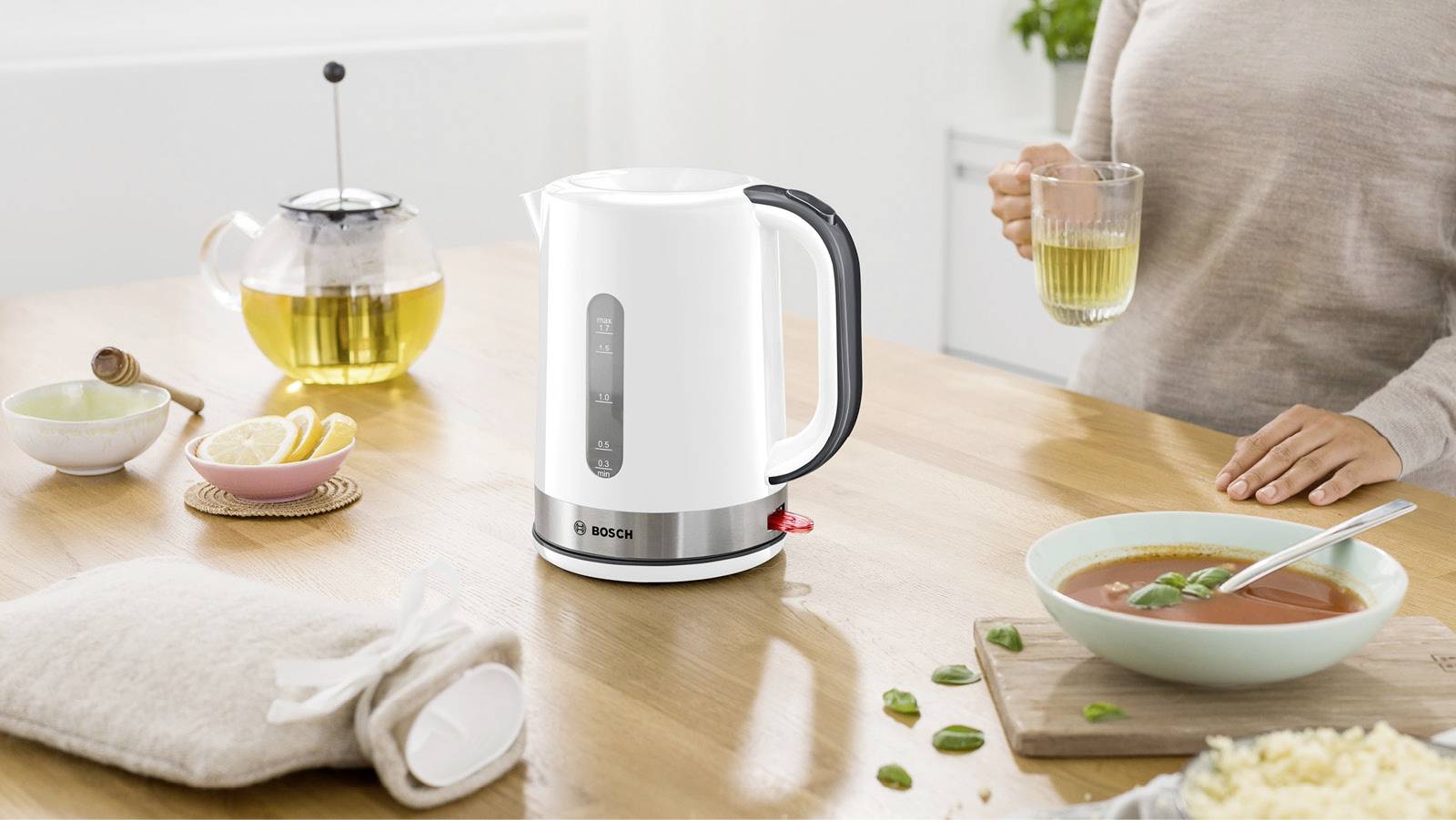 'White kettle on a wooden table, visually dominating the image. In the background, a teapot, lemons, and a person with a tea cup.'