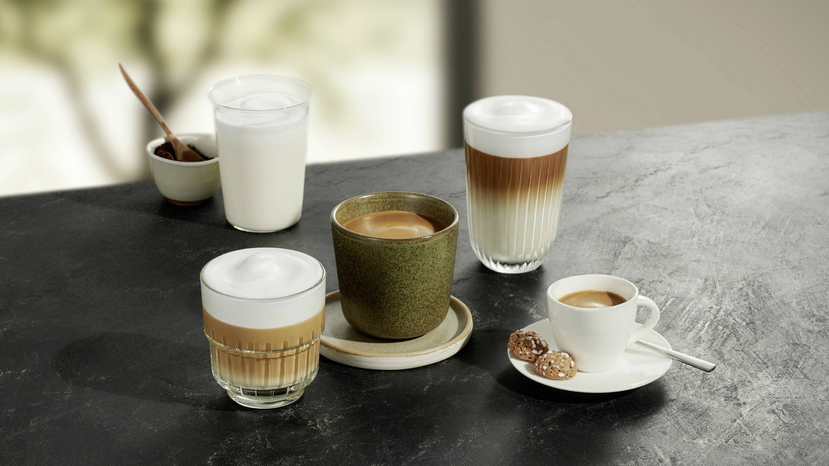 Five different coffee specialities and a glass of milk are sitting on a table; a colourful selection of beverages.