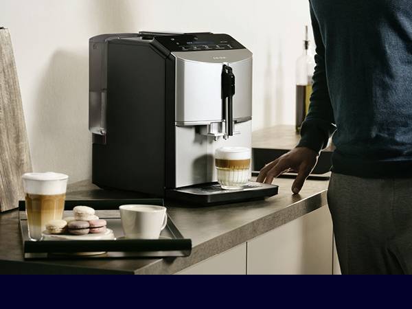A person is preparing coffee using a fully automatic coffee machine. On the table, there are two glasses of coffee and some macarons.
