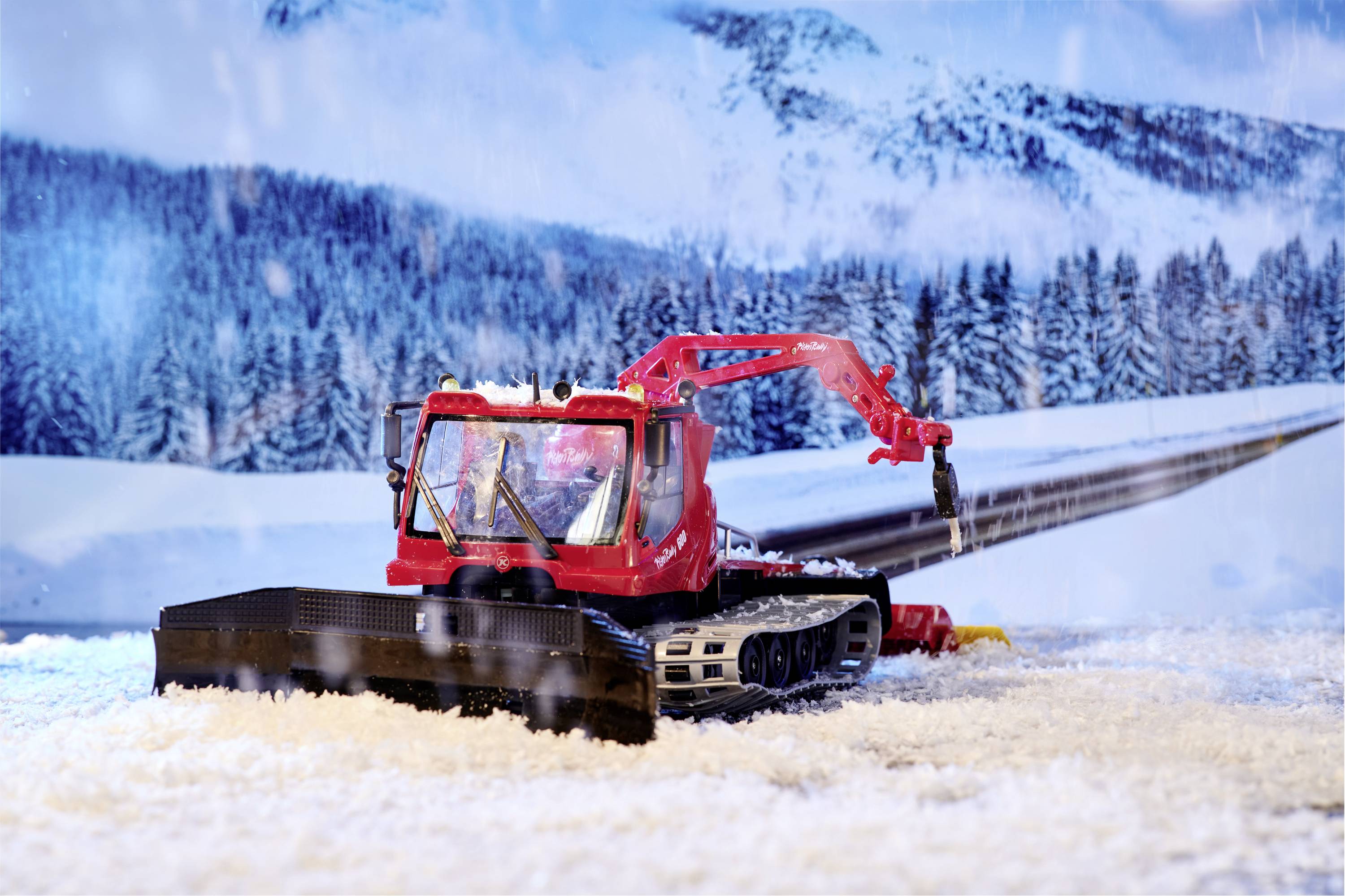 A snow groomer clears snow in a snowy mountain landscape with forested hills and mountains in the background.