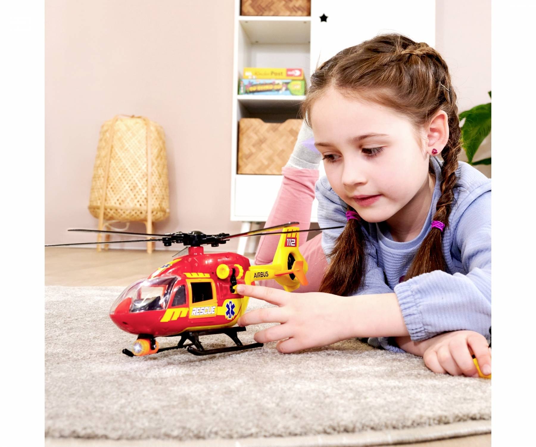 A girl is playing on a rug with a toy helicopter. In the background, there is a bookshelf with books and baskets.