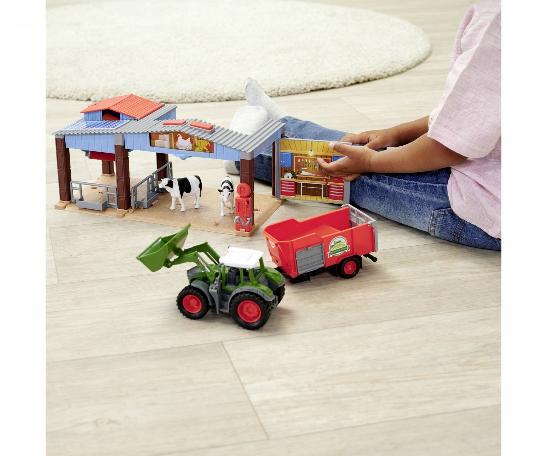 A child is playing with a toy farmyard. A barn, a cow figure, a tractor with trailer, and a play figure can be seen.