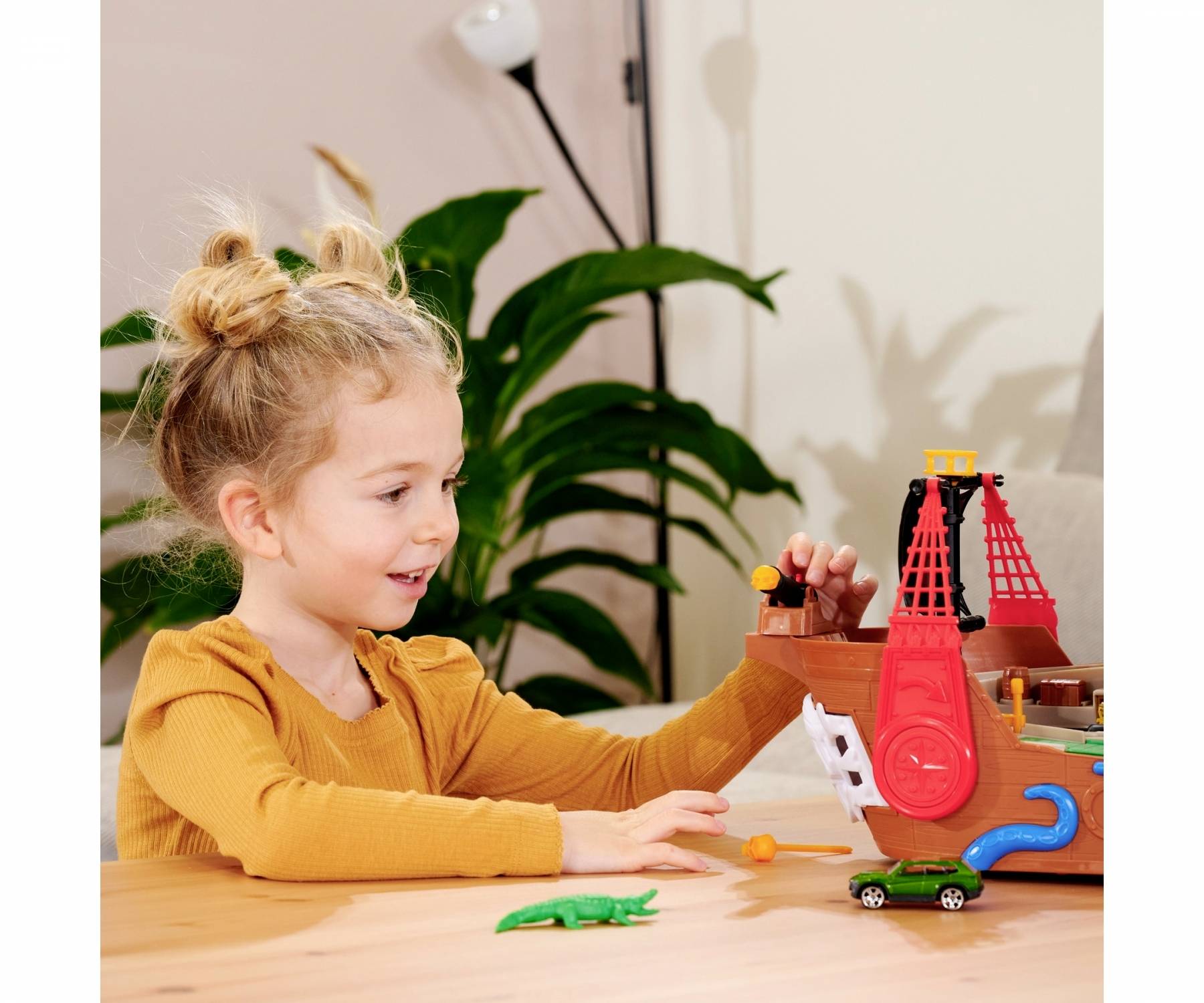 A child is happily playing with a colourful pirate ship toy on a table. A plant is visible in the background.
