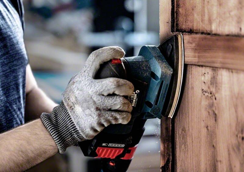 A person is sanding a wooden door with an electric sanding tool. They are wearing work gloves and focusing intently on the task.