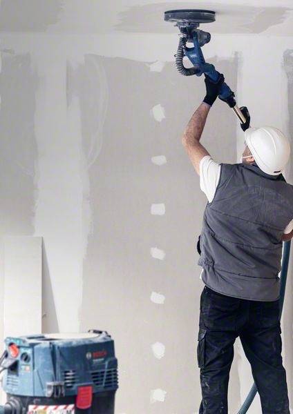 A construction worker wearing a hard hat and protective equipment is grinding a wall. Unfinished walls and tools can be seen in the background.