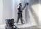 A worker wearing a hard hat and mask is smoothing a wall with a sanding machine in a room. A vacuum cleaner stands beside him.