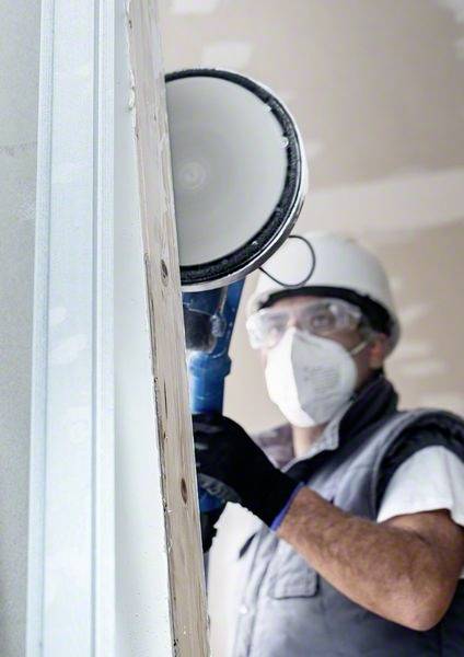 A construction worker wearing a hard hat and mask is grinding a wall with an electric sander inside a room.