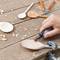 A hand is working on a wooden spoon with a Dremel tool on a wooden table. Wood shavings and additional spoons are visible.