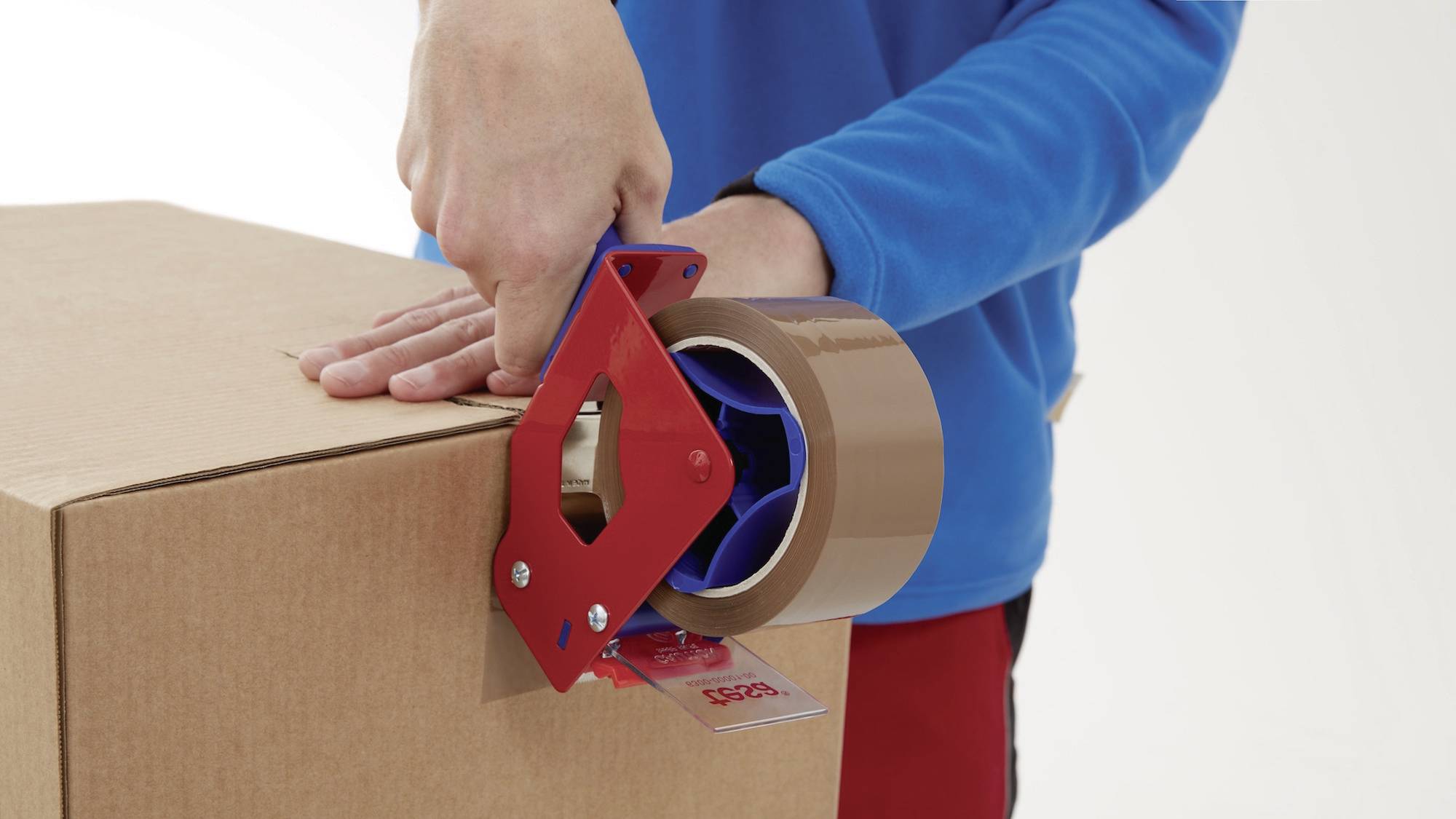 A person is sealing a cardboard box with brown parcel tape held in a red hand-held tape dispenser.