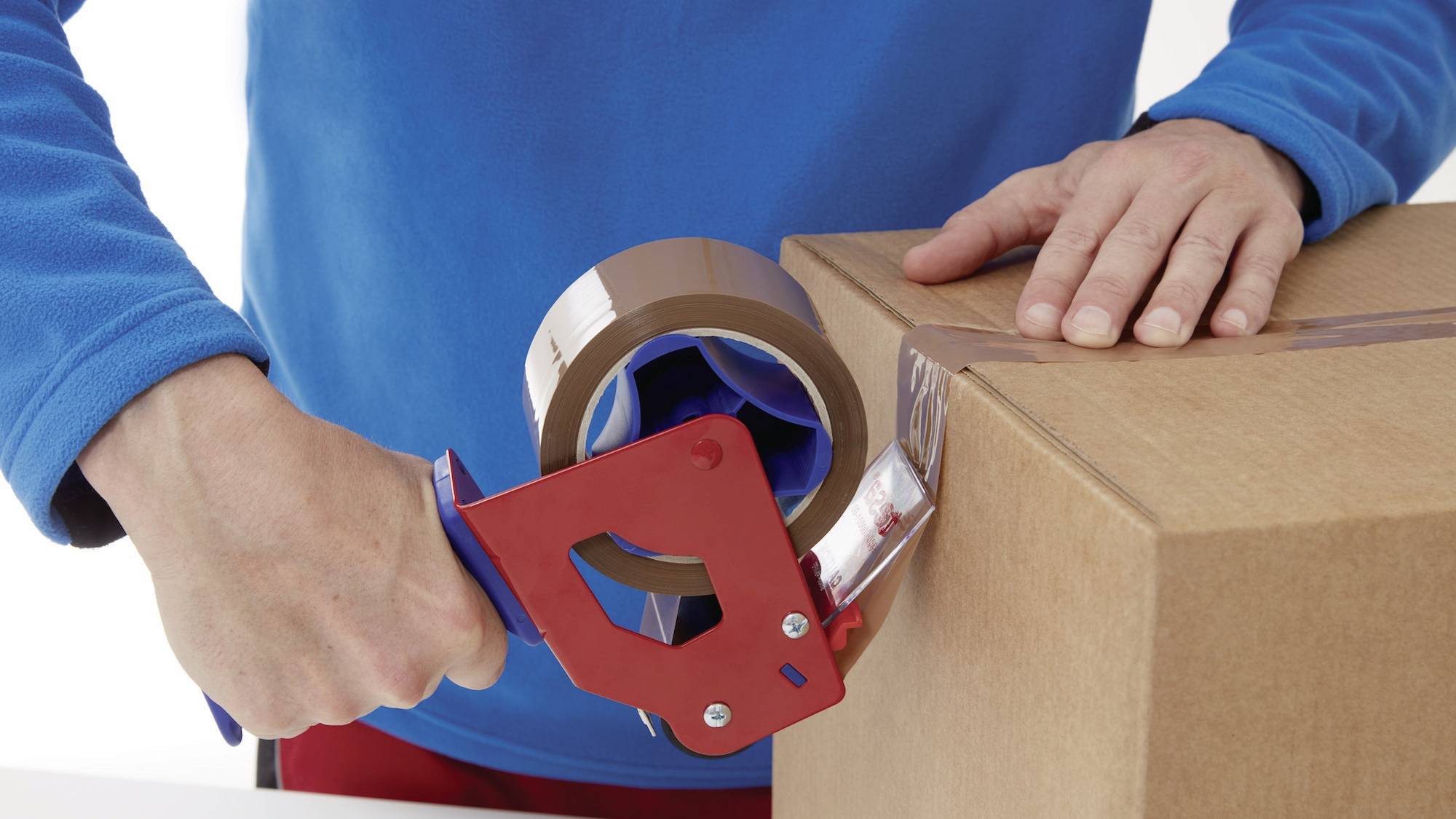 A person is sealing a cardboard box with a parcel tape dispenser. The box is resting on a work surface.