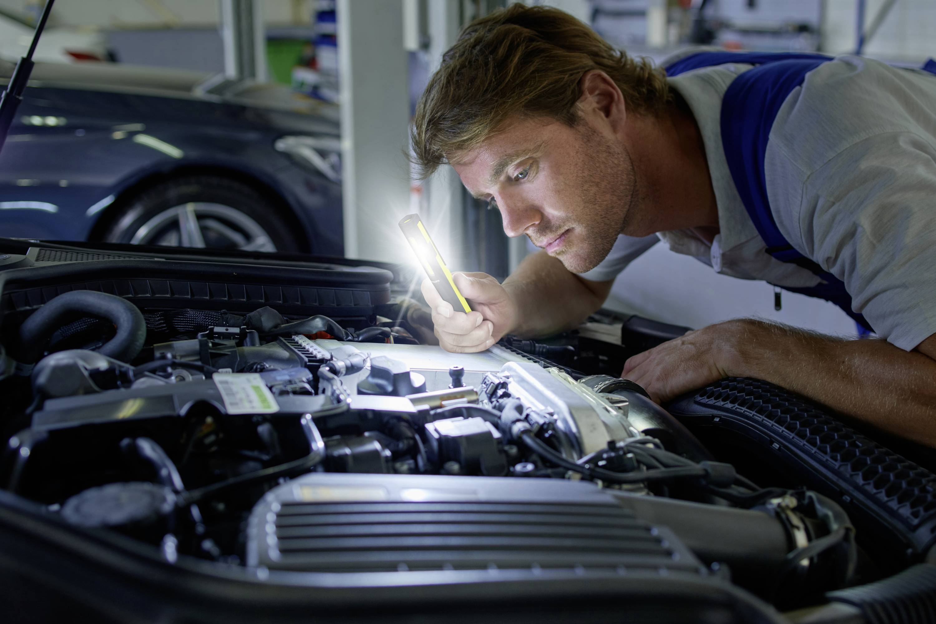 A mechanic inspects the car engine with a torch in a workshop. He is focusing intently on the engine details.