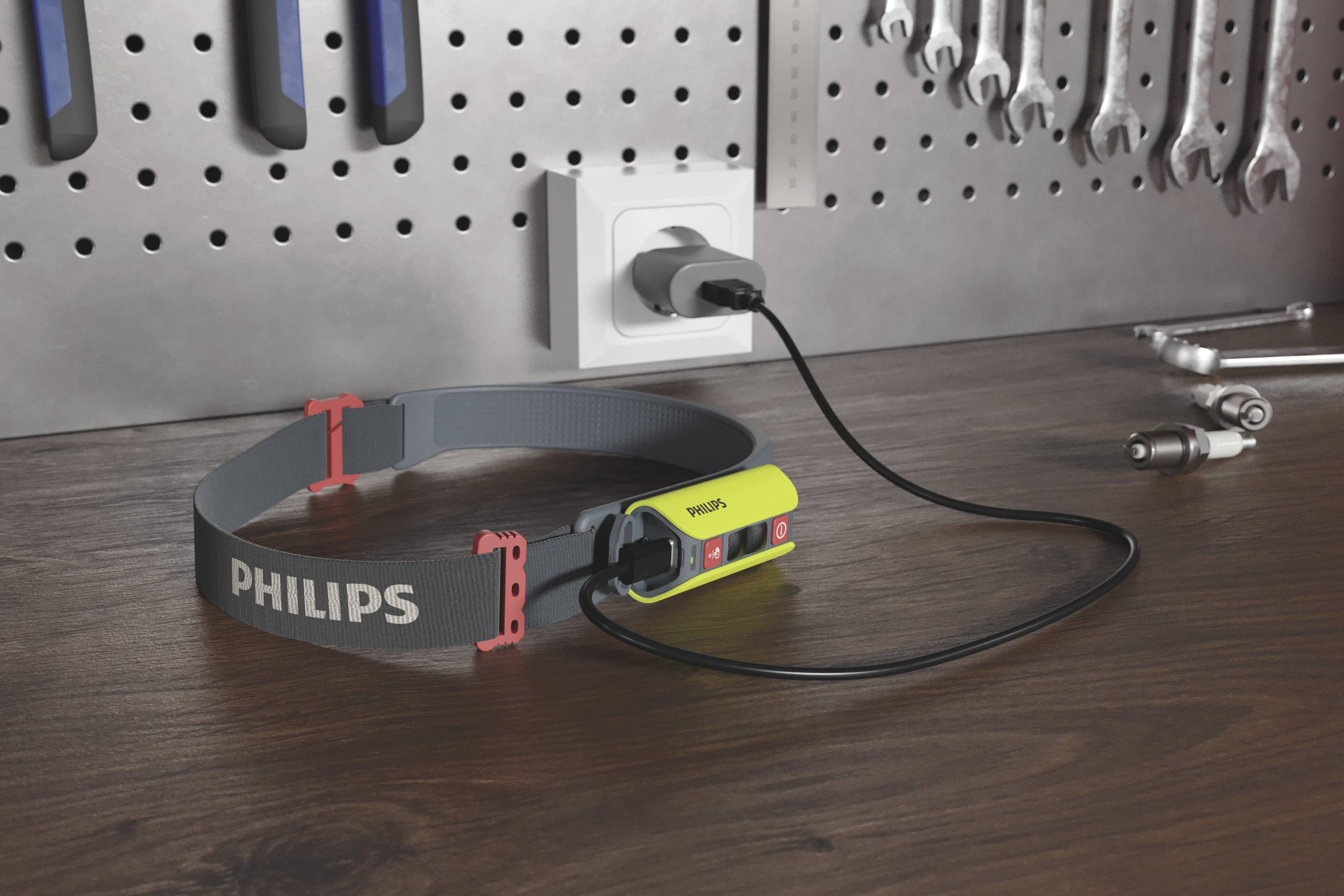 A Philips head torch lies on a workbench, plugged into a socket. In the background, tools hang on a wall.