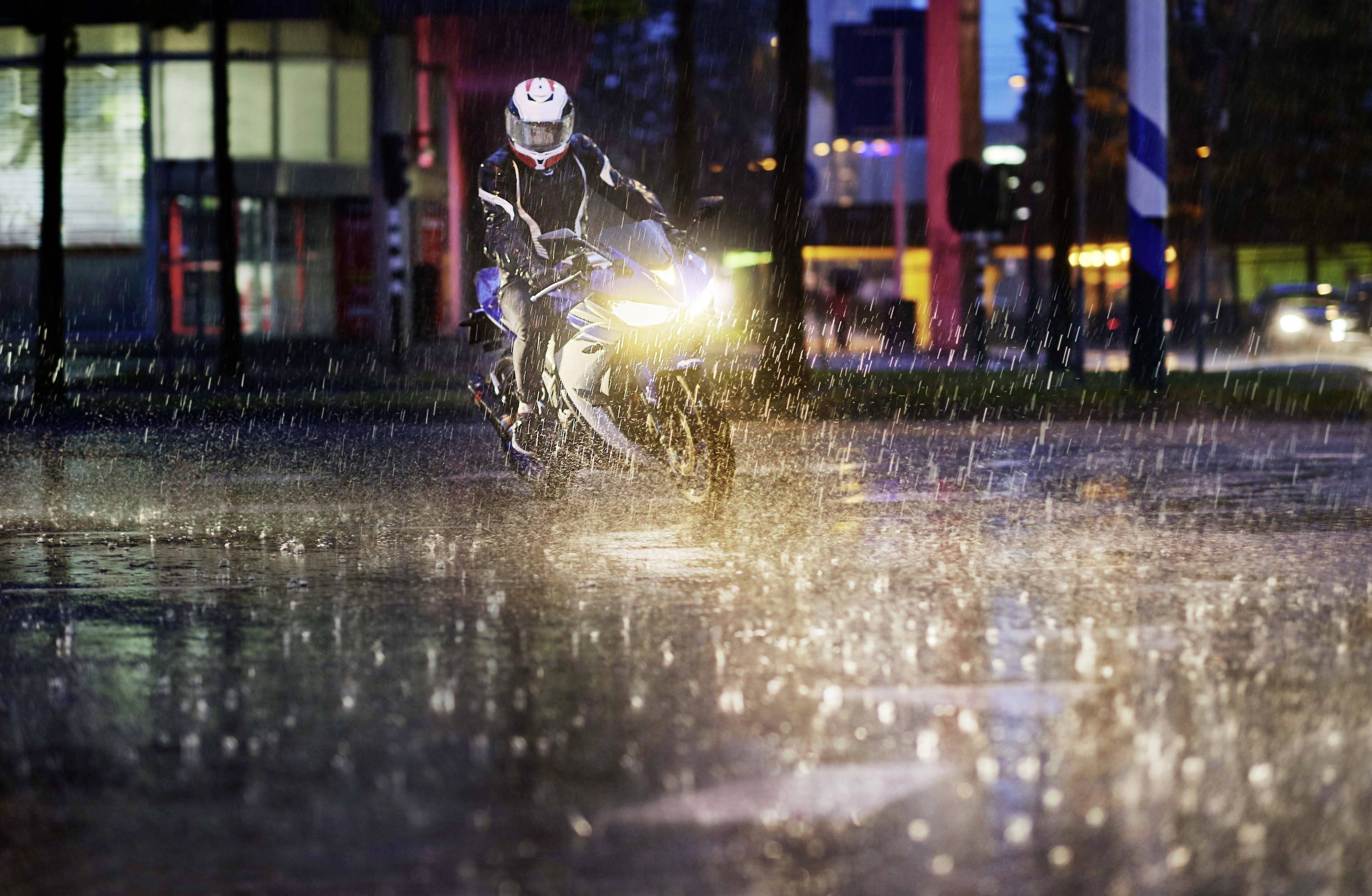 A person is riding a motorbike through a lit city street in heavy rain, with lights reflecting on the wet road surface.