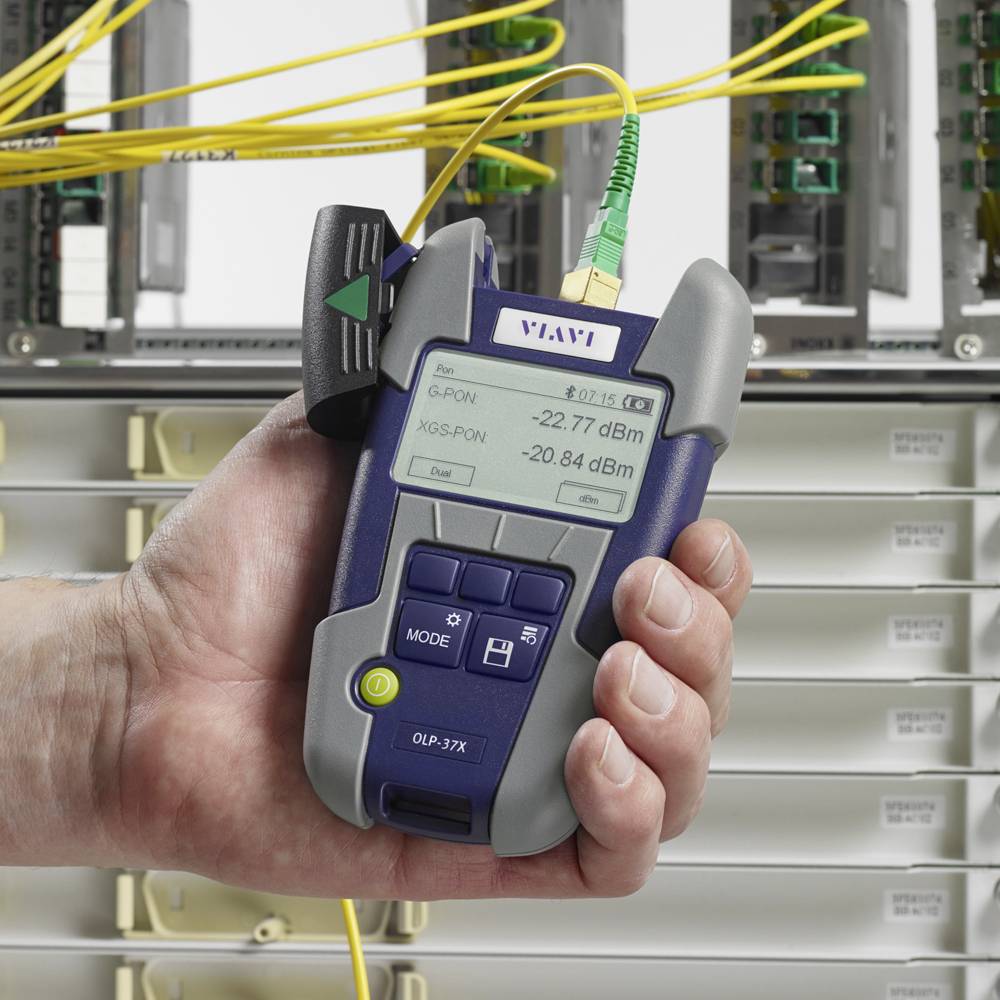 A hand holds a device for measuring optical fibre signals in front of a wired fibre optic distribution panel. The display shows power values.