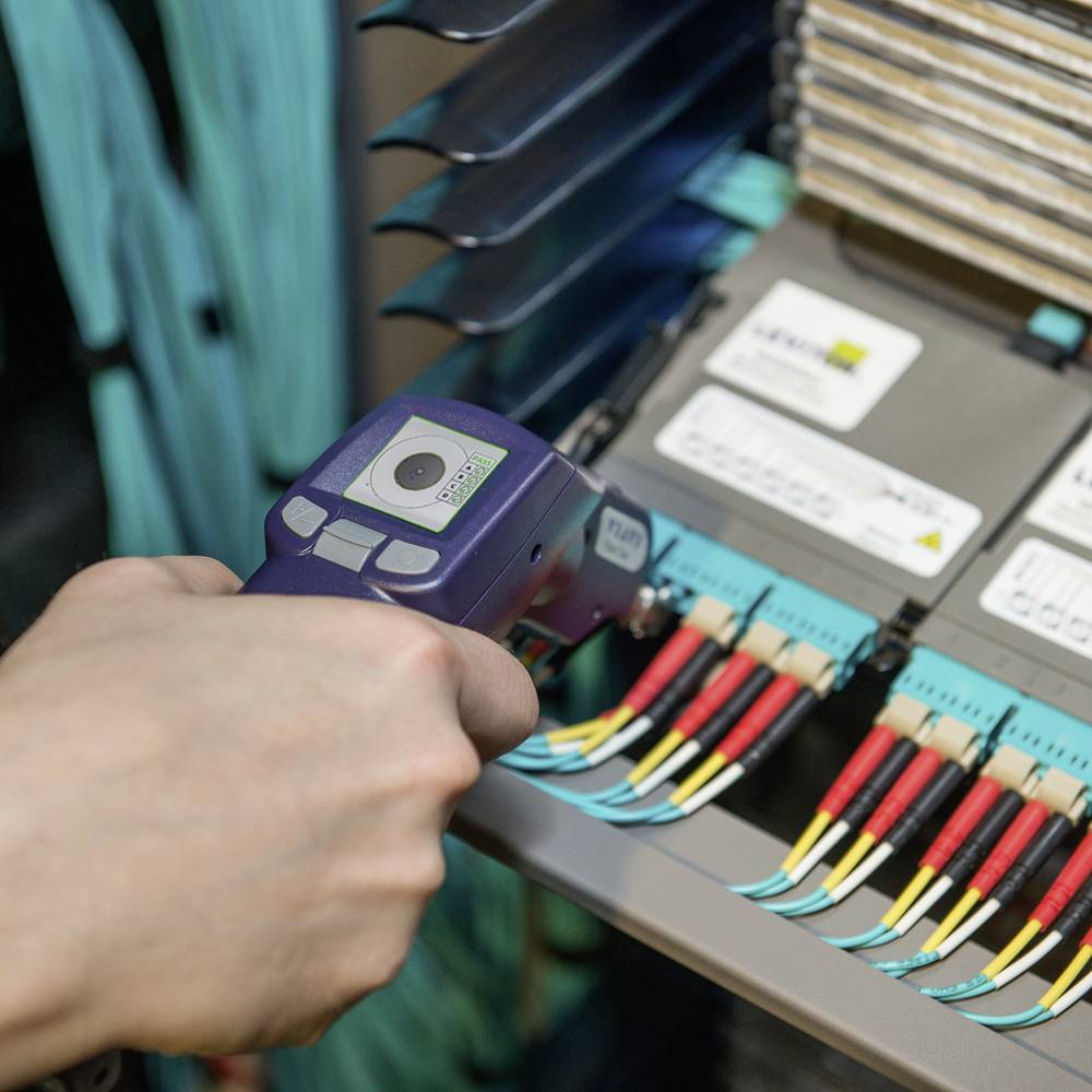 A hand holds a fibre optic testing device next to a cable connection panel with coloured cables. The environment appears technical and clean.