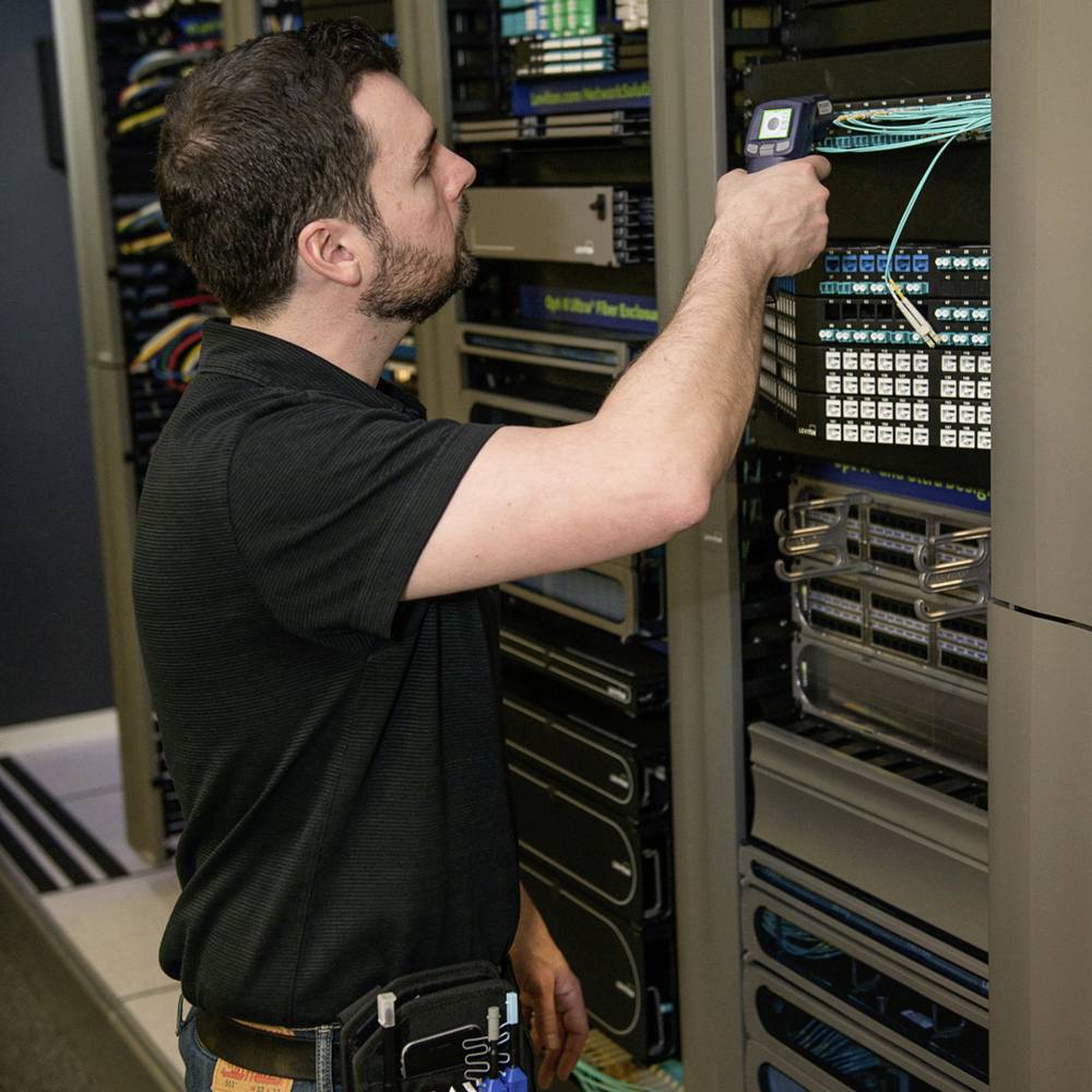 A technician is inspecting a network device in a server rack, surrounded by cables and other hardware, in an IT room.
