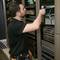 A technician is inspecting a network device in a server rack, surrounded by cables and other hardware, in an IT room.