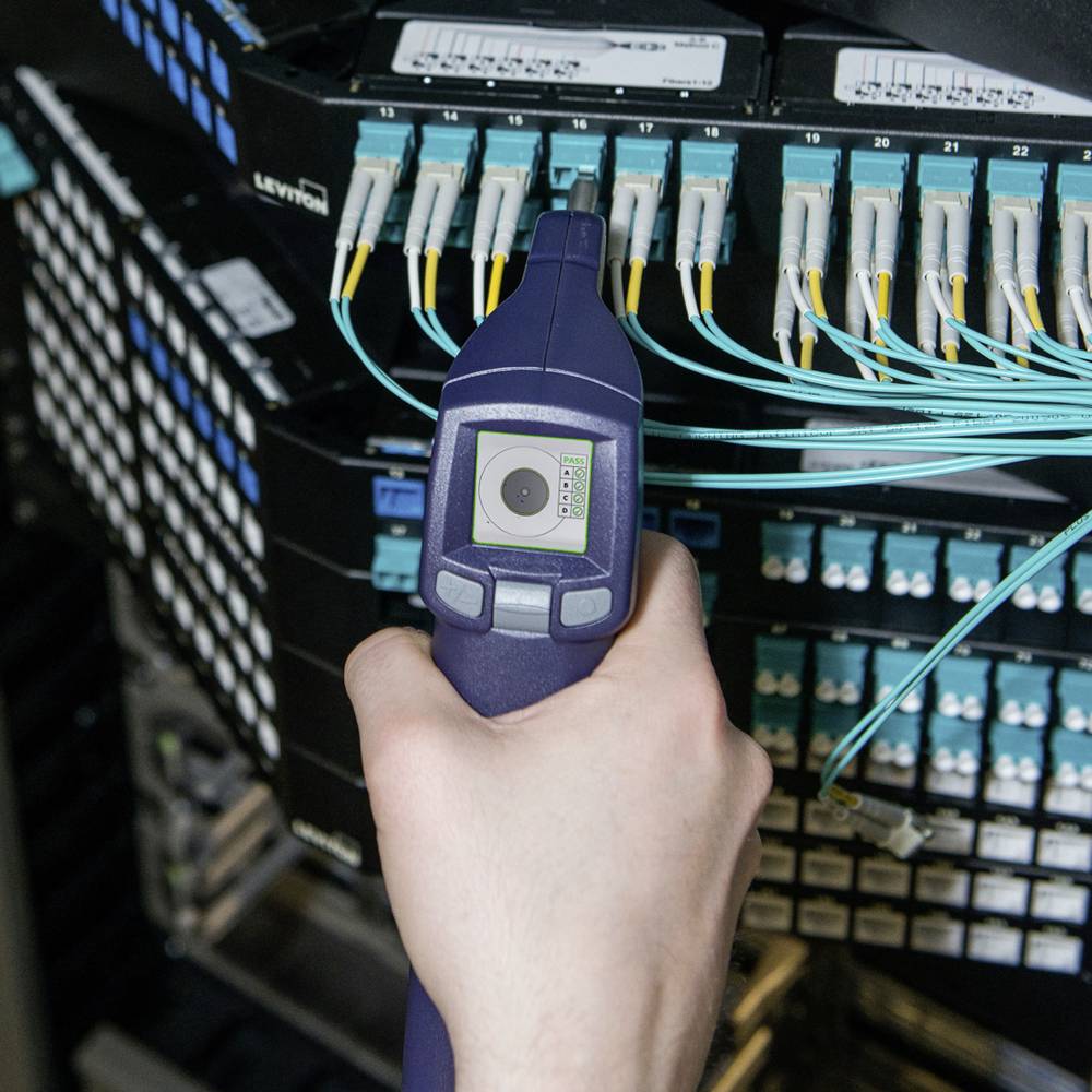 A hand is holding a testing device connected to a patch panel with numerous cable connections in order to test network connections.