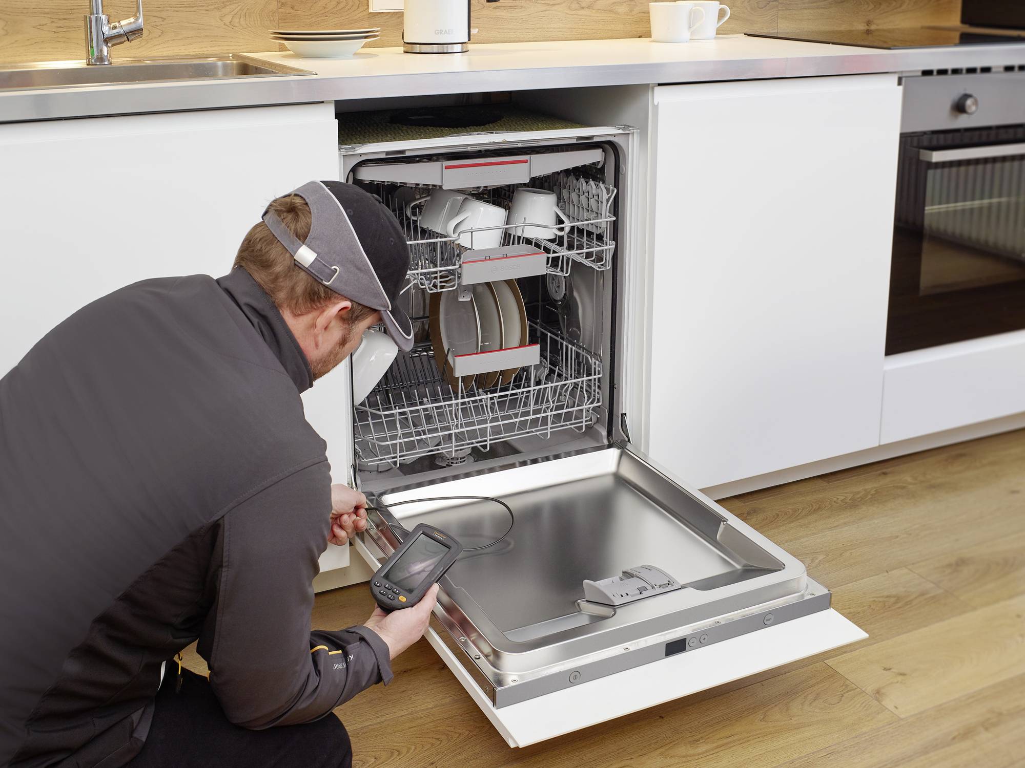 A technician is inspecting an open dishwasher in a modern kitchen. Crockery and cups can be seen inside the appliance.