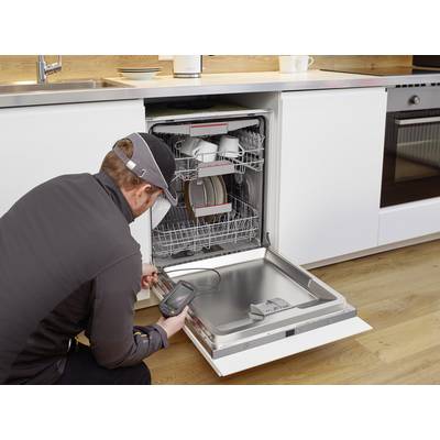A technician is inspecting an open dishwasher in a modern kitchen. Crockery and cups can be seen inside the appliance.