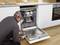 A technician is inspecting an open dishwasher in a modern kitchen. Crockery and cups can be seen inside the appliance.