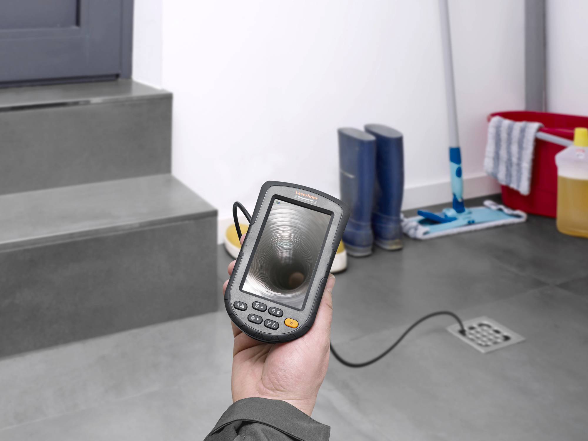 A person is holding an inspection device with a camera, showing the inner part of a pipe. Cleaning equipment and rubber boots are visible in the background.