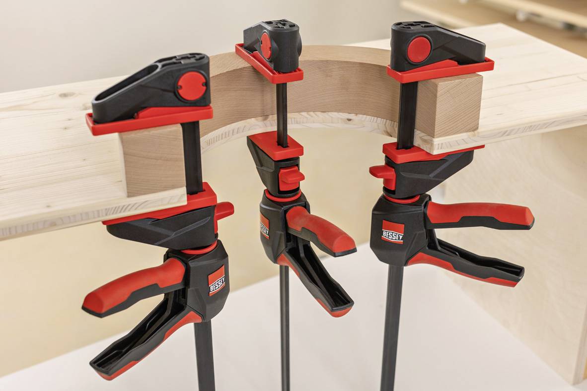 Three G-clamps are holding a curved piece of wood on a workbench. They are black and red and are made by BESSEY.