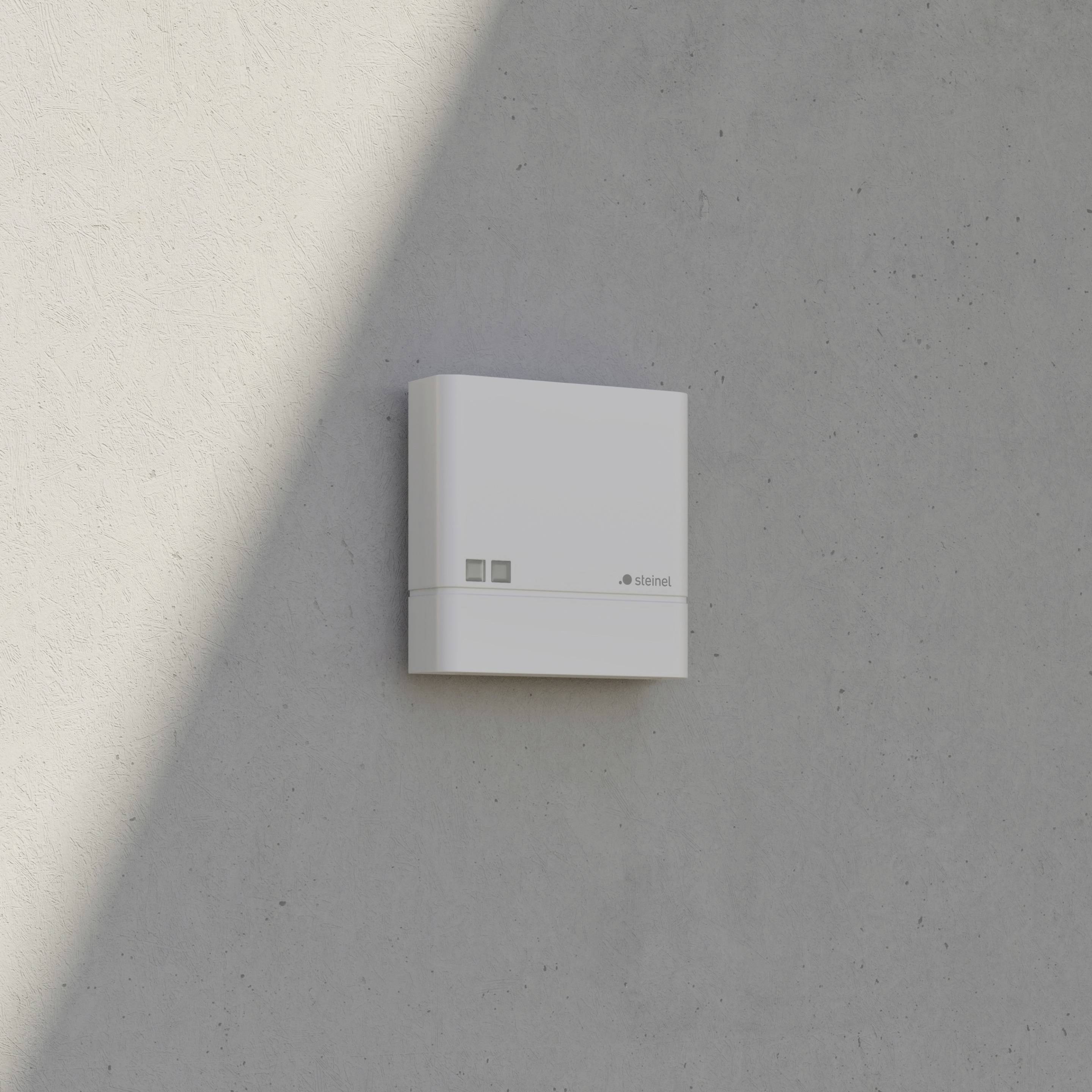 A white, square device mounted on a grey wall with two buttons. A shadow falls on the wall to the left.