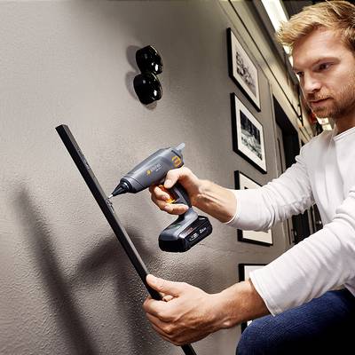 A man is drilling a hole in the wall with a cordless drill. Picture frames are hanging on the wall, and two round plug sockets are visible.