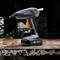 Cordless hot glue gun with an ergonomic grip, placed on a workbench. Blurred workshop environment in the background.