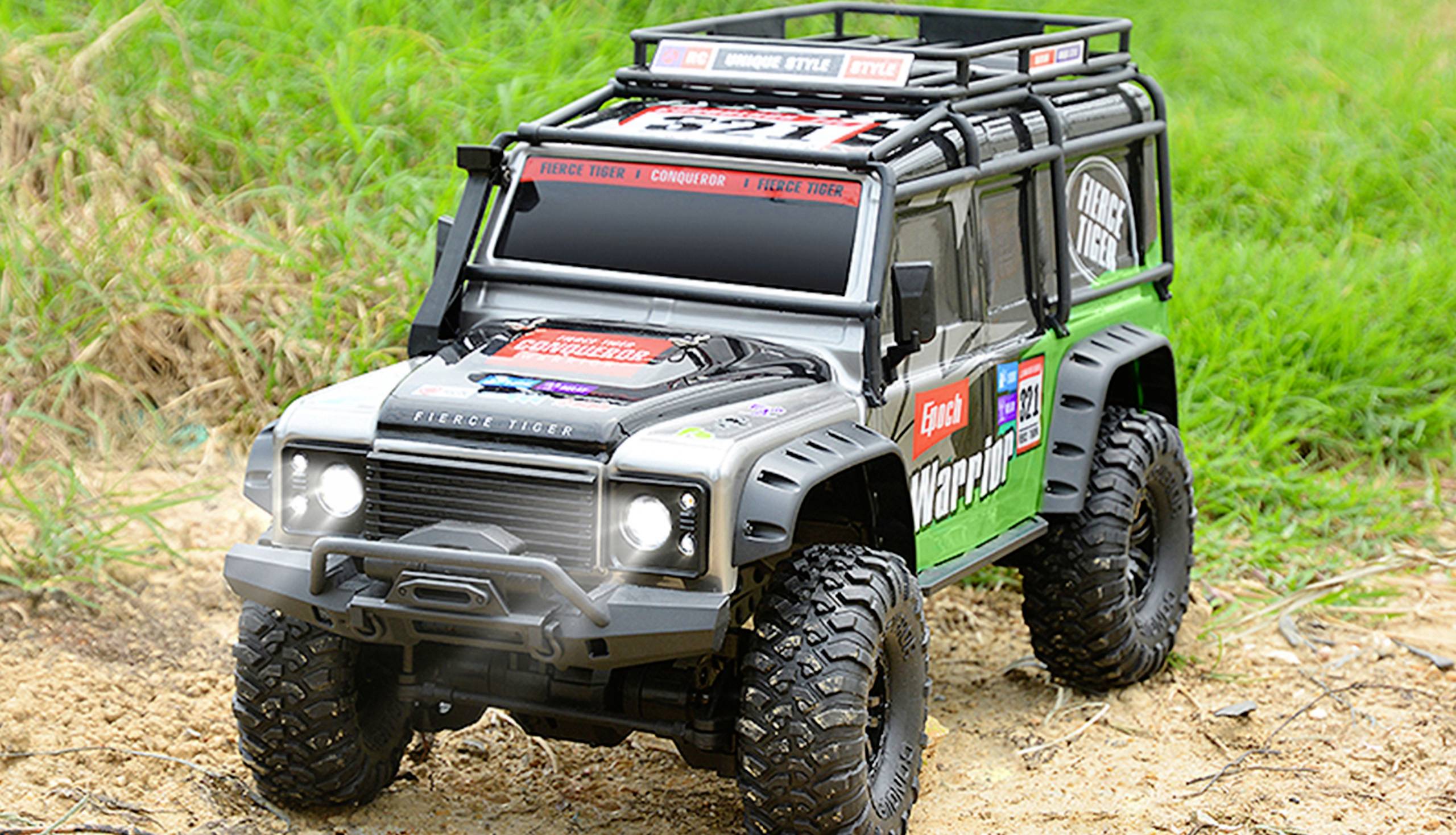 A model off-road vehicle drives over sandy earth and grass. It has large tyres and sporty stickers.